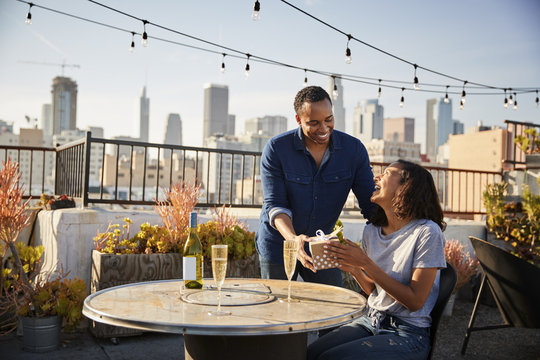 Man Giving Woman Gift As They Celebrate On Rooftop Terrace With City Skyline In Background