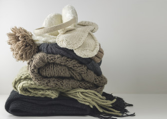 Warm woolen knitted winter and autumn clothes, folded in a pile on a white table. Sweaters, scarves, gloves, hat, headphones. Place for text. Copyspace.