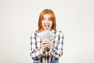 Young beautiful redhead woman screaming in rejoice, laughing w/ open mouth, fistful of one hundred dollar bills like fan. Excited attractive female with lots of cash. Background, copy space, close up.