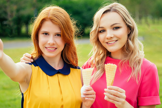 two girlfriends beautiful young ginger redhead woman in a yellow dress and blonde female person in a pink dress eating vanilla ice cream in a waffle cone in the summer park take picture selfie phone