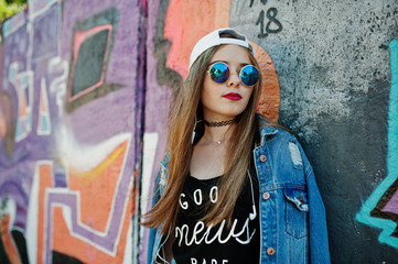 Stylish casual hipster girl in cap, sunglasses and jeans wear, listening music from headphones of mobile phone against large graffiti wall.