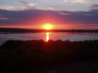 red-yellow sun setting behind a forest and reflected in a river against a cloudy sky background