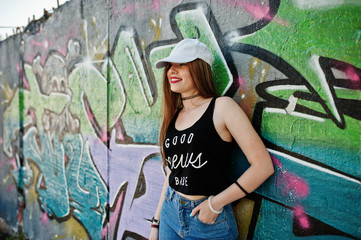 Stylish casual hipster girl in cap  and jeans wear against large graffiti wall.
