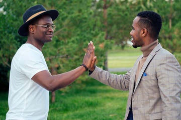 two black men in stylish suits meeting in a summer park. African-Americans friends hispanic businessman embrace high five greeting each other teamwork outdoors. successful deal concept