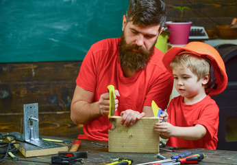 Father, parent with beard teaching little son to sawing while son play with toy saw. Boy, child busy in protective helmet learning to use handsaw with dad. Educational games concept.