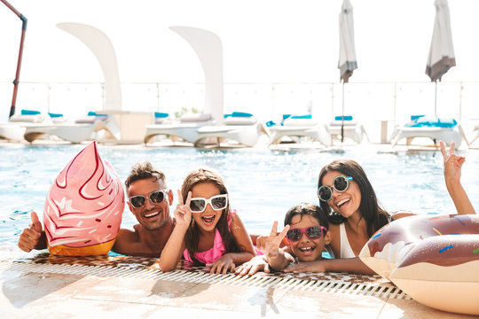 Photo of modern cheerful family with children wearing sunglasses swimming in pool, with rubber ring outside hotel