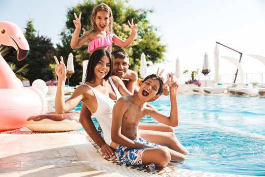 Image of attractive caucasian family with children sitting near luxury swimming pool, with pink rubber ring outside hotel