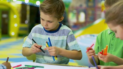 Group of interested young children of elementary school age sitting at table in play center and drawing with colored felt tip pens