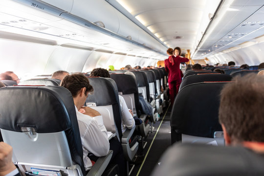 Interior of commercial airplane with unrecognizable passengers on their seats during flight. Stewardess in red uniform walking the aisle of commercial airplane.