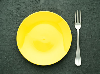 Blank flat yellow plate, fork on dark gray stone concrete table, top view. Mock up, copy space