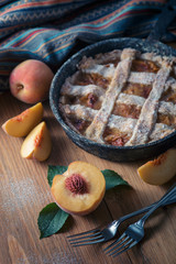 Peach pie in a cast-iron frying pan lies on a wooden table. On the table lie cut peaches, green leaves, fabric, knife and forks.