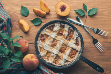 Peach pie in a cast-iron frying pan lies on a wooden table. On the table lie cut peaches, green leaves, fabric, knife and forks.