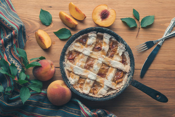 Peach pie in a cast-iron frying pan lies on a wooden table. On the table lie cut peaches, green leaves, fabric, knife and forks.