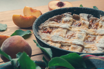 Peach pie in a cast-iron frying pan lies on a wooden table. On the table lie cut peaches, green leaves, fabric, knife and forks.