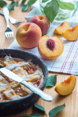 Peach pie in a cast-iron frying pan lies on a wooden table. On the table lie cut peaches, green leaves, fabric, knife and forks.