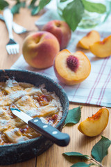 Peach pie in a cast-iron frying pan lies on a wooden table. On the table lie cut peaches, green leaves, fabric, knife and forks.