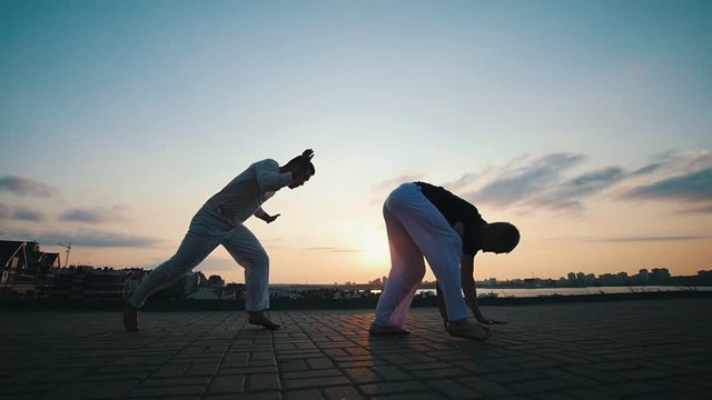 Strong men train, dance capoeira on the asphalt over the city, against the beautiful sky