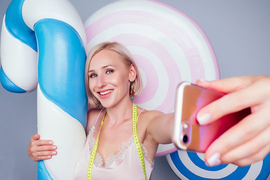 beautiful young blonde Barbie sweet woman confectioner sexy model perfect makeup hug a huge lollipop with measuring tape and take pictures selfie on phone background fake sweets candy in studio shot