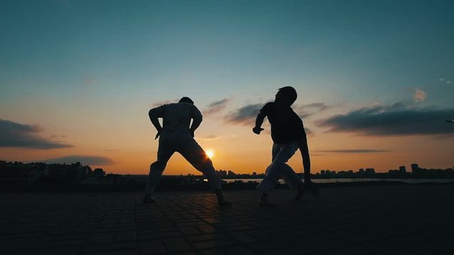 Silhouettes of two sports men who train skills of martial art of capoeira against the background of the amazing sky
