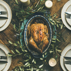 Flat-lay of whole roasted chicken in tray for Christmas eve celebration, plates, glasses and candles over rustic wooden background, top view, square crop. Holiday table decoration set concept