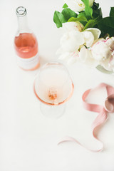 Rose wine in glass and bottle, pink decorative ribbon, peony flowers over white background, copy space. Summer celebration, wedding greeting card, invitation concept