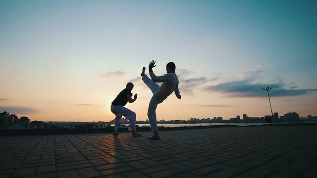 Men with a sporty physique dancing capoeira outdoors, on the pavement, against the beautiful promenade and summer sunset