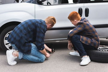father lifting car with floor jack for changing tire, son sitting on tire
