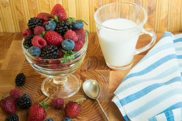 Berry raspberries strawberry blueberries in a glass vase on a wooden set of bars. A useful berry breakfast on a wooden table with a striped blue and white towel, a silver spoon and a glass of milk