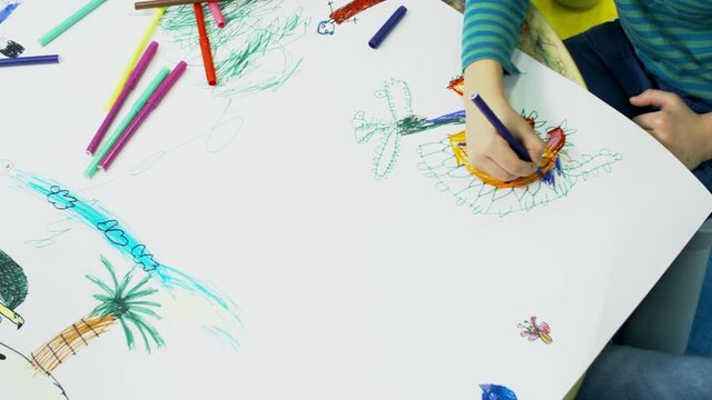 Tracking shot of children of primary school age using colored felt tip pens when drawing pictures on big sheet of paper, above view