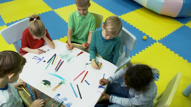 High angle view of multi ethnic group of little children sitting at table in bright playroom and drawing beautiful picture together using colored felt tip pens