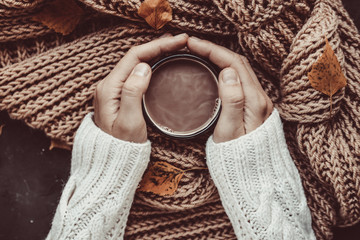 A cup of warm cocoa in female hands on a background of brown, knitted scarf and autumn leaves. A...