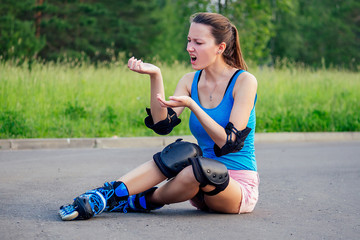 attractive young athletic slim brunette woman in pink shorts and blue top with protection elbow pads and knee pads on roller skates sitting on the asphalt in the park . fall concept