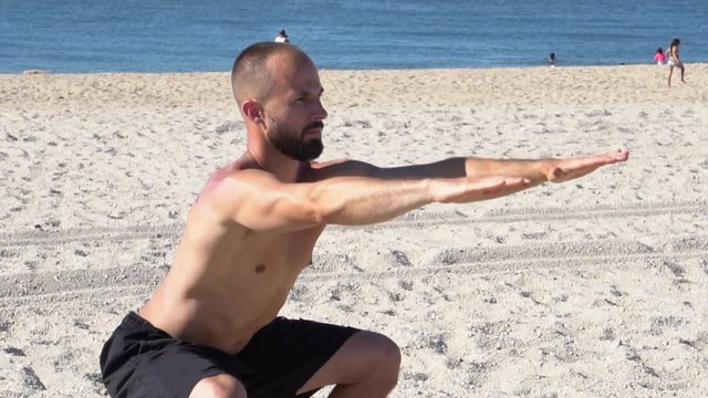 Young athletic build man exercising on beach waterfront with ocean in background. Free standing squats to strengthen core muscles on sand without shirt