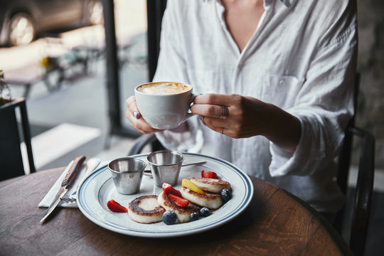 cropped shot of woman in white shirt holding cup of coffee at restaurant above syrniki on table