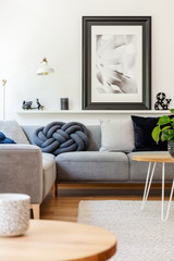 Poster above grey sofa with cushions in white living room interior with lamp and table. Real photo
