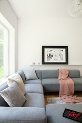 Pink blanket and pillows on grey corner couch in white living room interior with poster. Real photo