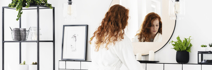 Ginger haired woman getting ready in the morning in white bathroom interior with lamps, mirror and poster placed on wall shelf