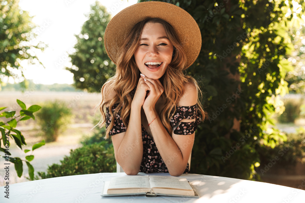 Poster Cheerful laughing young woman sitting in cafe outdors in park with book.