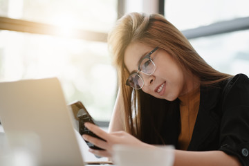 Young woman sitting in coffee shop at wooden table, drinking coffee and using smartphone.On table is laptop. Girl browsing internet, chatting, blogging. Female holding phone and looking on his screen.