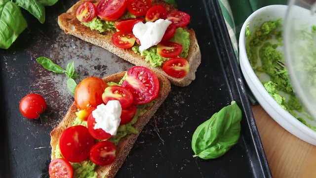A woman places burrata cheese and fresh basil leaves on avocado toast and cherry tomatoes