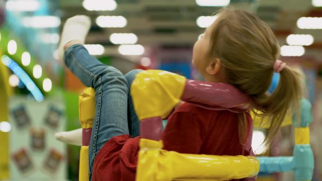 Medium shot of cute little girl of elementary school age spinning in chair when riding merry go round in children play center