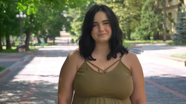 Beautiful young woman with obesity smiling and looking at camera, standing on street in park, charming and happy