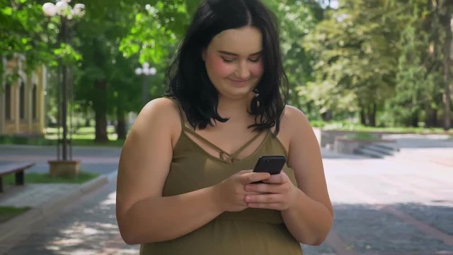 Beautiful obese brunette woman typing on phone and standing on street in park near university, sunny day outside