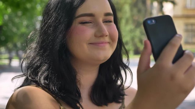 Portrait of young beautiful woman with obesity taking selfie with her phone, smiling and standing on street in park, happy
