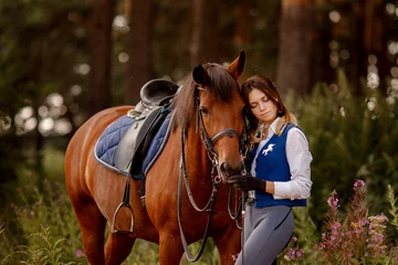 Foto op Canvas Young woman rider equestrian stands next to brown horse in forest. © Parilov