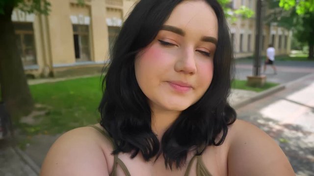 Charming young happy woman with obesity taking selfie or recording video and smiling, standing on street near college building