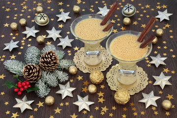 Christmas eggnog drink in glasses with gold star and bell decorations, foil wrapped chocolate balls with winter flora of holly, fir and pine cones on rustic oak table background. Festive theme.