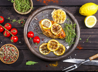 Grilled chicken fillet with fresh thyme, cherry tomato, lemon, garlic and spices in a slate plate on black background