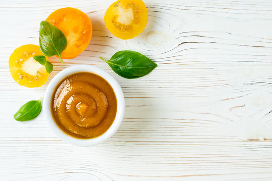 Tomato ketchup sauce in a bowl with basil and yellow tomatoes on white wooden table. Yellow ketchup. Top view. Copy space