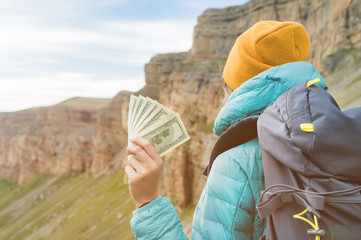 A traveler girl wearing a hat and sunglasses is holding a hundred dollar bills in the hands of a fan against the backdrop of cliffs on nature. Back view